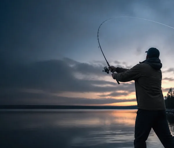 Fiskare i motljus som kastar med haspelspö från stranden i skymning, med spöet böjt och linan på väg ut över vattnet.