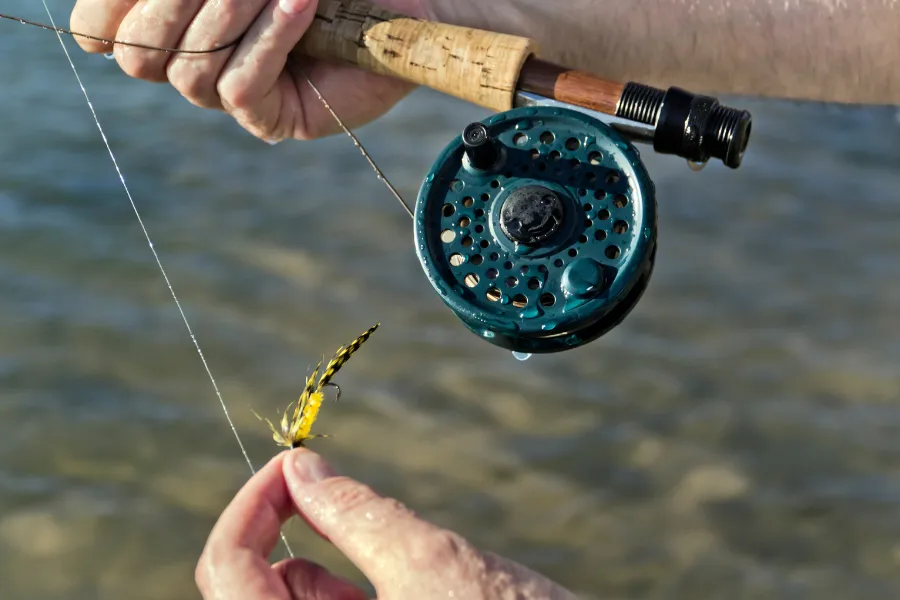 Närbild av flugfiske – fiskare håller ett flugspö med rulle och en gul fluga över vattnet.
